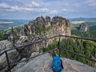 Blauer Rucksack mit Wanderstöcken auf felsigem Aussichtspunkt mit Blick auf Schrammsteine-Felsformationen mit Wäldern und Tälern im Hintergrund.