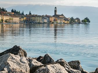 View of Salò town on Lake Garda with historic buildings and church tower reflected in calm water. Rocky shore in foreground, mountains in background.