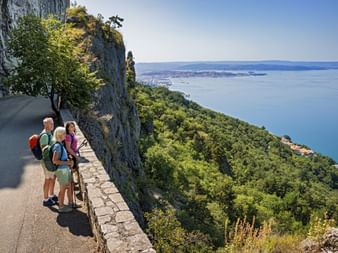 Drei Wanderer mit Rucksäcken stehen an steinerner Aussichtsmauer mit Blick auf Adria und Triester Küste vom Julische Alpen Höhenweg.