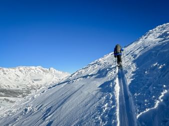 Skitourengeher mit Rucksack steigt einen steilen verschneiten Hang in Maria Alm-Dienten hinauf, unter klarem blauen Himmel mit schneebedeckten Bergen.