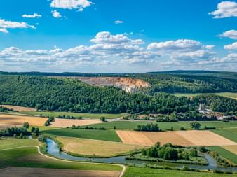 Panoramic view of Altmühltal valley with limestone cliffs, winding river, patchwork fields, and dense forests under blue sky with white clouds.