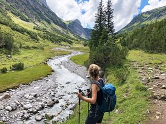 Wanderin mit blauem Rucksack und Wanderstöcken betrachtet Gebirgsbach im grünen Alpental am Lechweg, Österreich.