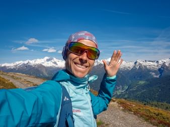 Lächelnde Wanderin in türkiser Jacke und Skibrille macht Selfie am Eggerjoch mit schneebedeckten Alpengipfeln im Hintergrund unter blauem Himmel.
