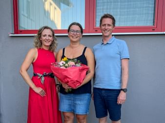 Drei Personen im Freien: Frau in rotem Kleid, Bettina in der Mitte mit Blumenstrauß und Mann in blauem Poloshirt.