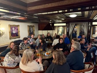 Group dining in elegant ship restaurant with dark wood beams, classical portraits, and round tables. People enjoying drinks and conversation.