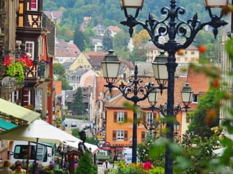 Verzierte schwarze Straßenlaterne im Vordergrund mit Blick auf eine historische Straße im Elsass. Bunte Fachwerkhäuser und rote Ziegeldächer sichtbar.