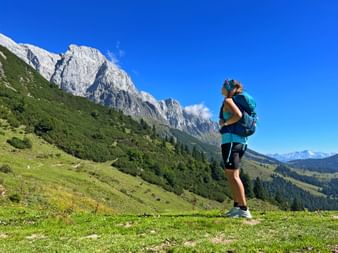 Female hiker with backpack standing on green alpine meadow, viewing dramatic limestone peaks in the Bavarian and Salzburg Alps under blue sky.