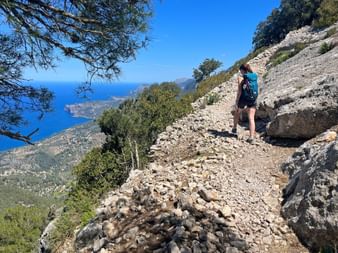Female hiker with backpack walking on rocky GR221 mountain trail in Mallorca with Mediterranean Sea and coastal landscape visible below.