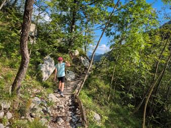 Girl with red cap hiking uphill on rocky forest trail with green trees and blue sky. Mountain view visible through the foliage.