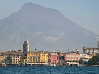 Colorful historic buildings of Riva del Garda along Lake Garda waterfront with a prominent clock tower and dramatic mountain rising in the background.
