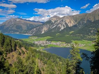 Aerial view of Reschensee and Haidersee lakes surrounded by mountains. A village nestles between the lakes with green meadows and forests.