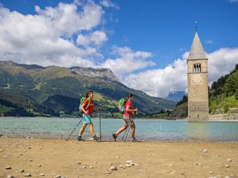 Hikers on the shore of Lake Reschen with a view of the church tower