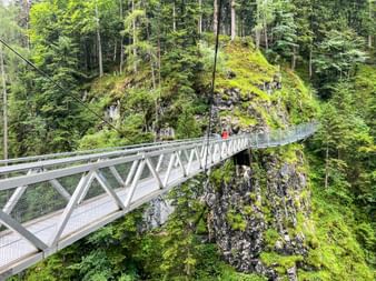Metallene Hängebrücke über eine moosbedeckte Felsschlucht in der Geisterklamm. Dichter Wald umgibt die Brücke mit einem Wanderer in Rot.