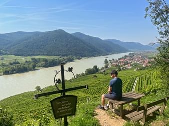 Hiker resting on wooden bench overlooking Danube River, vineyards, and Weissenkirchen village in Wachau Valley with forested mountains.