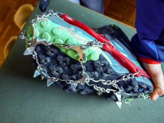 Nahaufnahme eines Wanderstiefels mit Grödel aus Metallketten an der Sohle für Wintertraktionen. Der Stiefel steht auf einer grünen Schaumstoffmatte.