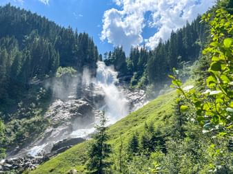 Oberer Krimmler Wasserfall fließt über felsige Hänge in Hohe Tauern, umgeben von grünen Wiesen und bewaldeten Bergen unter blauem Himmel.