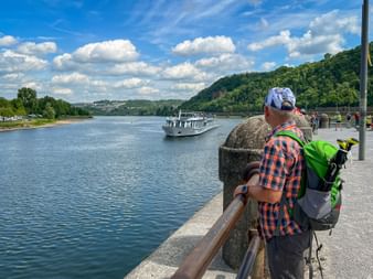 Wanderer mit Rucksack beobachtet ein Passagierschiff auf dem Rhein am Deutschen Eck in Koblenz, mit grünen Hügeln und blauem Himmel im Hintergrund.