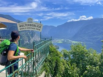 Wanderin mit grünem Rucksack am Welterbeblick mit Aussicht auf See und Berge des Salzkammerguts unter blauem Himmel.