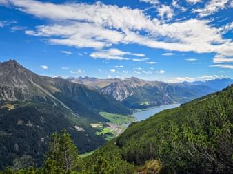 Panoramic view of Reschensee lake nestled in an Alpine valley, surrounded by forested mountain slopes and distant peaks under a blue sky.