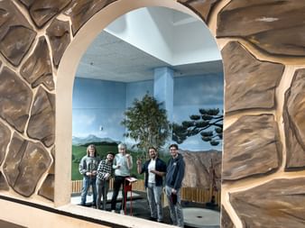 Fünf Teammitglieder stehen in einem gewölbten Durchgang mit Steinwandrahmen auf einer Indoor-Minigolfanlage mit gemalten Landschaftsbildern.