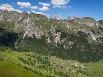 Green alpine valley with scattered trees and mountain huts near Vallmingalm. Rocky peaks with green slopes rise under blue sky with clouds.