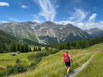 Wanderin mit rotem Rucksack auf Bergweg in Obernberg. Grüne Wiesen, Wälder und dramatische Alpengipfel unter blauem Himmel.