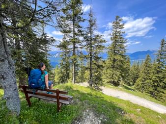 Wanderin mit blauem Rucksack sitzt auf Holzbank umgeben von Nadelbäumen und blickt auf Bergtal mit fernen Gipfeln.