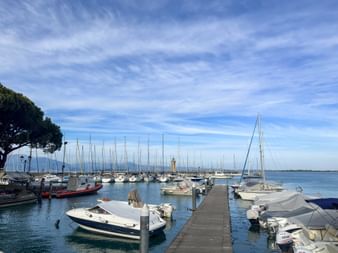 Hafen in Padenghe sul Garda mit weißen Booten am Holzsteg. Gelber Leuchtturm sichtbar, Berge im Hintergrund unter blauem Himmel.