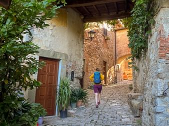 Wanderer mit blauem Rucksack in Kopfsteingasse in Montefioralle, Toskana. Steingebäude mit Efeu, Topfpflanzen und Straßenlaterne sichtbar.