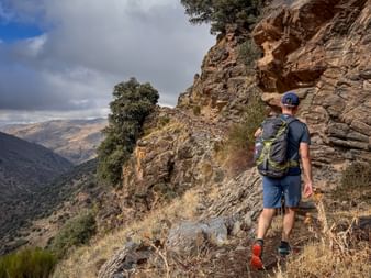 Wanderer mit Rucksack auf schmalem felsigen Pfad entlang einer Felswand in Alpujarras, mit Bergtal und bewölktem Himmel im Hintergrund.