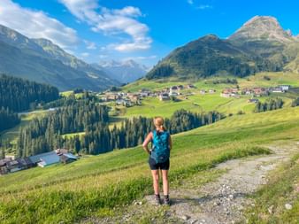 Female hiker with blue backpack standing on grassy hillside overlooking Warth village in the Allgäu Alps with mountains and blue sky.