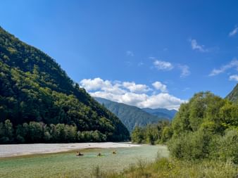 Drei Kajakfahrer auf der türkisfarbenen Soča mit bewaldeten Berghängen und Kiesbänken in den Julischen Alpen unter blauem Himmel.
