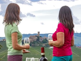 Zwei Frauen mit Weingläsern blicken auf das Dorf Barolo und Weinberge in Langhe, Piemont. Hügelige Landschaft mit Weinreben und historischen Gebäuden.