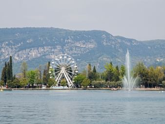 White ferris wheel on the shore of Lake Garda in Bardolino with water fountain, trees, and mountains in the background under a hazy sky.