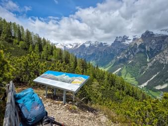 Blauer Rucksack neben Panoramatafel an Bergaussichtspunkt bei Gossensass, mit schneebedeckten Gipfeln und grünem Tal.