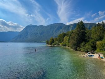Lake Bohinj with turquoise water, sandy beach with swimmers, forested shoreline, and Julian Alps mountains under blue sky with clouds.