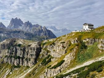 Auronzohütte auf einem grünen Alpenkamm mit den dramatischen Drei Zinnen in den Dolomiten hinter felsigem Gelände unter bewölktem Himmel.