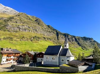 White church with dark roof and steeple in Pfelders village, surrounded by traditional alpine buildings and green mountain slopes under blue sky.