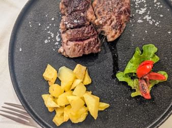 Sliced grilled steak with coarse salt on a dark plate, served with roasted potatoes and fresh salad with red peppers.