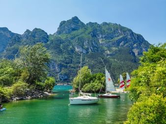 Sailboats moored in turquoise waters at Riva del Garda with dramatic mountain peaks in the background and lush green trees framing the scene.