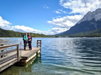 Zwei Wanderer stehen auf einem Holzsteg am Eibsee mit klarem türkisfarbenem Wasser, bewaldeten Ufern und der Zugspitze im Hintergrund.