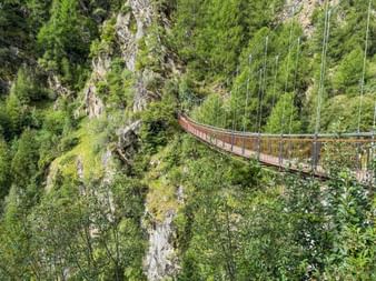 Wooden suspension bridge spanning a steep gorge with lush green vegetation and rocky cliffs on the Meraner Höhenweg hiking trail.