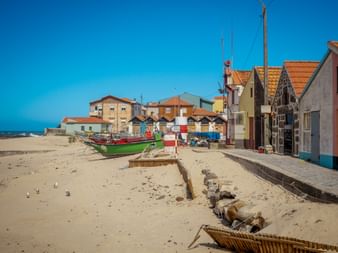 Bunte Fischerboote am Sandstrand in Vila Chã mit traditionellen Fischerhäusern mit orangefarbenen Ziegeldächern unter blauem Himmel.