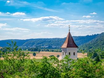 Weißer Kirchturm mit rotem Kegeldach im Altmühltal, umgeben von grüner Vegetation, Feldern und bewaldeten Hügeln unter blauem Himmel.