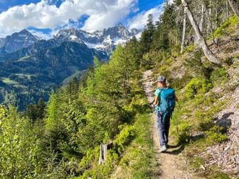 Wanderin mit Rucksack und Stöcken auf Bergweg im Hollersbachtal, Hohe Tauern. Schneebedeckte Gipfel und grüne Wälder im Hintergrund sichtbar.