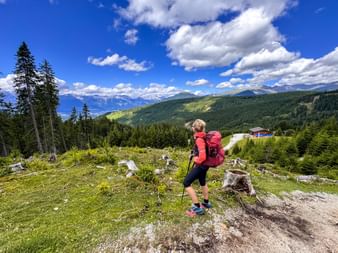Wanderin mit pinkem Rucksack und Wanderstöcken auf Bergweg bei Mieders, mit Karwendelgebirge und Wäldern im Hintergrund.