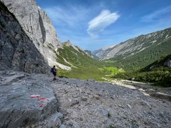 Wanderin auf felsigem Weg im Karwendelgebirge mit dramatischen Kalksteingipfeln, grünem Tal und Wald unter blauem Himmel.