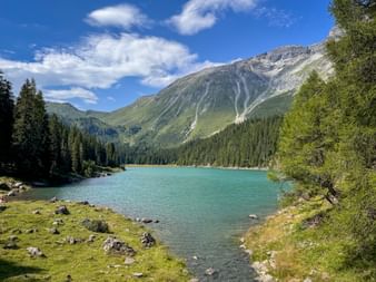 Türkisfarbener Obernberger See umgeben von Nadelwald mit dramatischen Berggipfeln im Hintergrund unter blauem Himmel mit weißen Wolken.