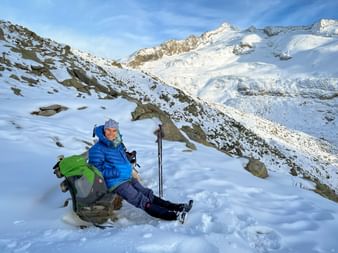 Lächelnde Wanderin in blauer Jacke sitzt im Schnee mit Rucksack und Stöcken, schneebedeckte Grossvenediger-Gipfel im Hintergrund.