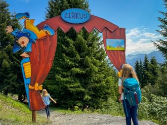 Colorful circus-themed entrance gate with clown figure and 'ZIRKUS' sign. Two children with backpacks stand before it on a mountain trail.